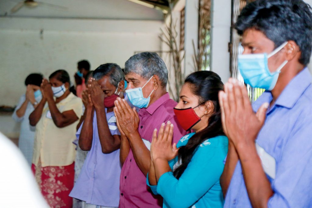 People stood in a line, some wearing masks. with their hands in a prayer position in front of their body